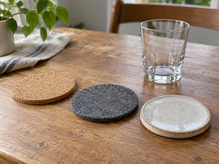 A cork coaster, felt coaster and glazed ceramic coaster side by side with a plain glass nearby, surface textures visible.