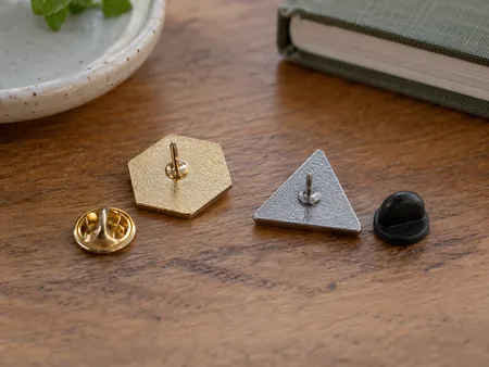 Back of two small original geometric metal enamel pins on a table, one with a butterfly clutch and one with a rubber back, backings separated.
