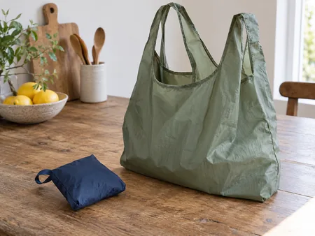 A thin reusable fabric tote folded into its attached small pouch beside the same style unfurled reusable bag in a different colour.