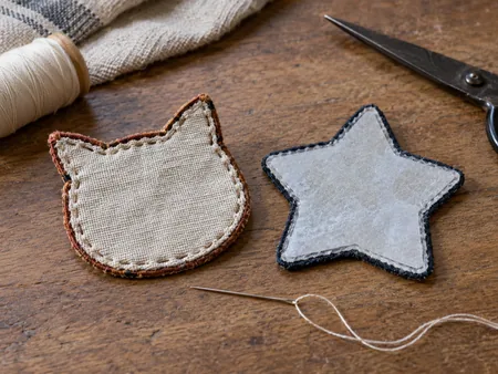 Two original plain embroidered fabric patches shown from back, one sew-on cloth backing and one iron-on adhesive backing, needle and thread beside them.