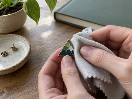 A small original geometric enamel pin gently wiped with a soft dry cloth on a tabletop, no liquid polish or abrasive tools.