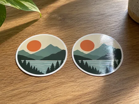 Two identical original geometric stickers side by side in raking light, one matte and one shiny gloss, reflected highlights only on gloss.