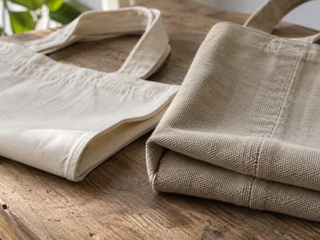 Two close folded edges of canvas totes of lighter and heavier cloth, dense woven canvas texture and stitching visible.